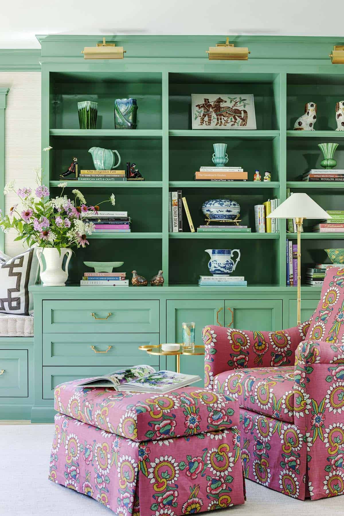 Glass-front green built-in bookshelves styled with vases, books, and pink floral armchair and ottoman
