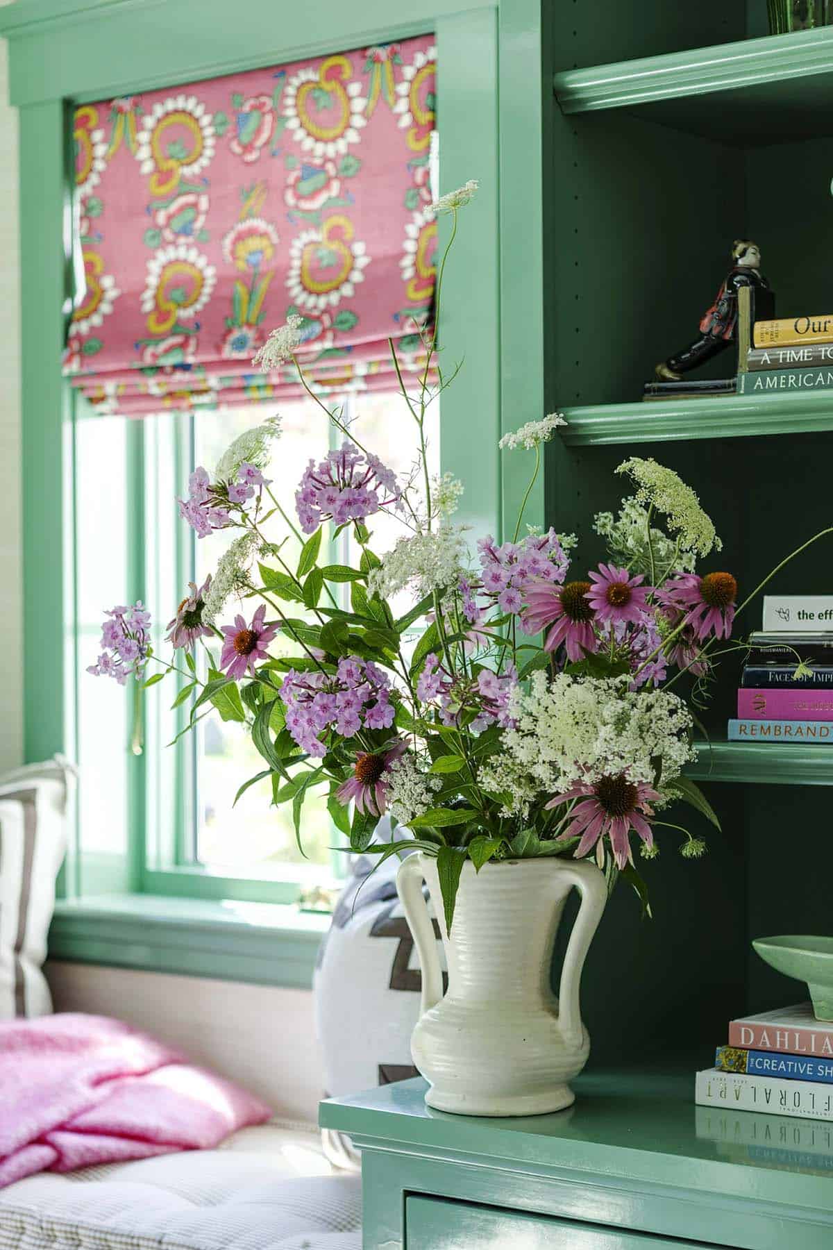 Close-up of wildflower bouquet in white pitcher beside green built-ins and a pink floral roman shade