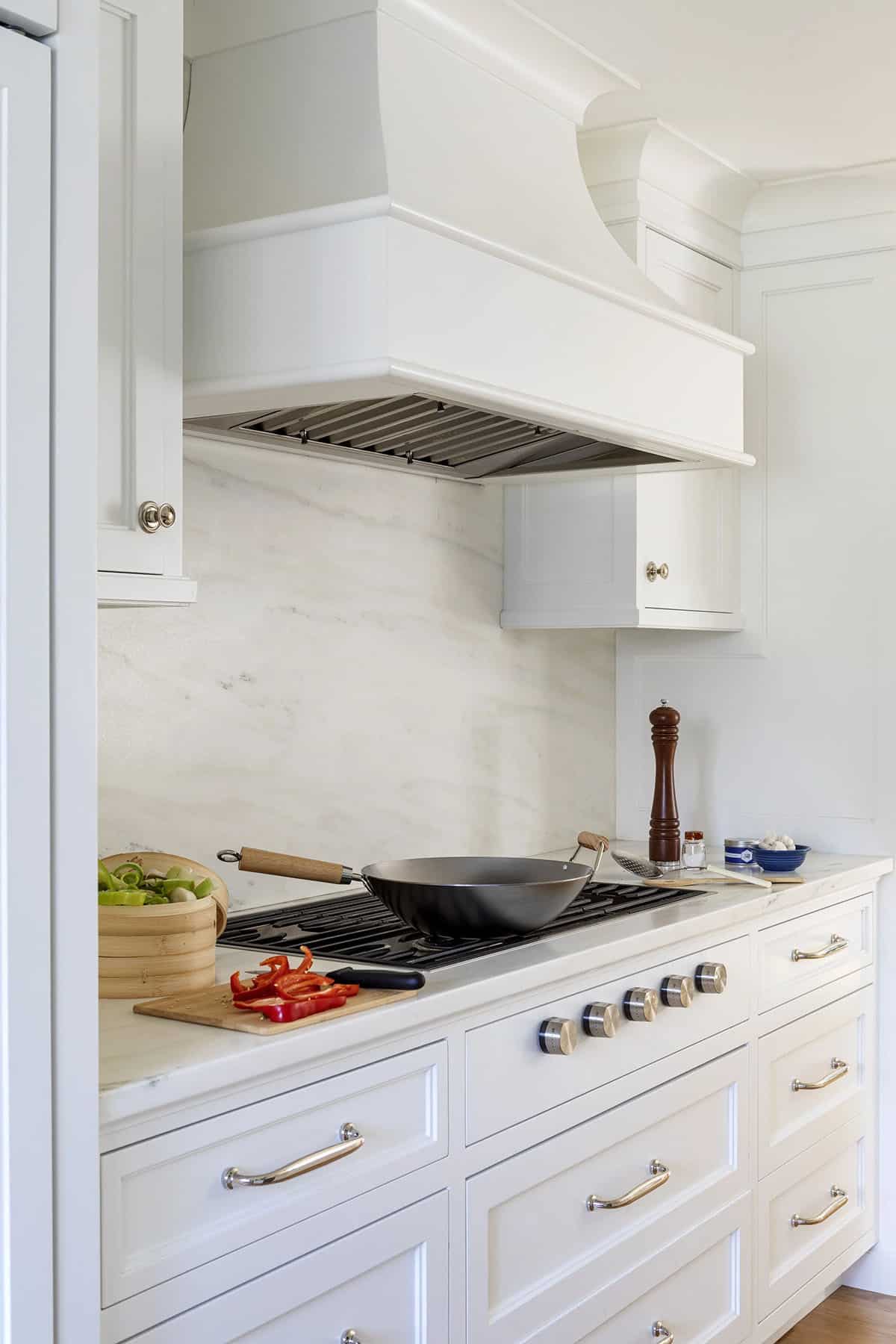 Close-up of white kitchen range with wok, marble backsplash, and brass-knobbed cabinetry