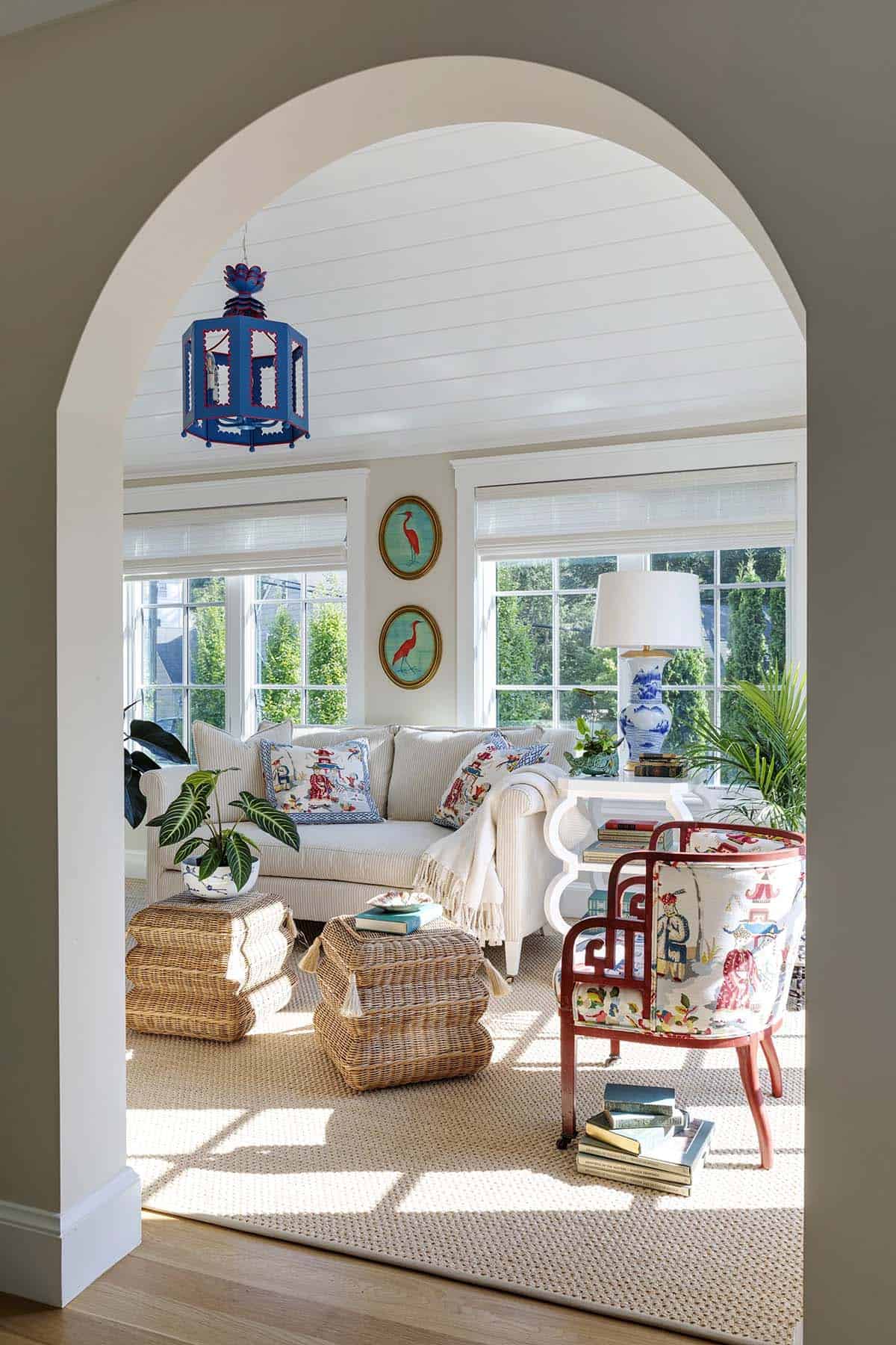 Archway view into a sunroom with striped sofa, rattan ottomans, and a red chinoiserie chair