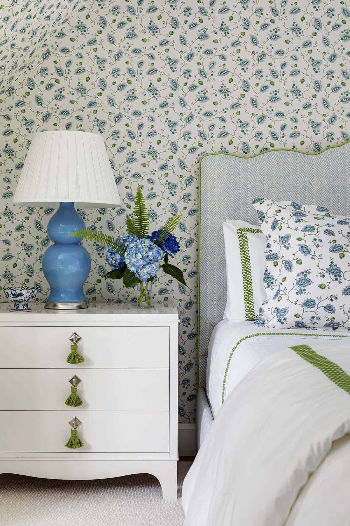 Bedside vignette with blue gourd lamp, white tasseled dresser, and blue-and-green floral wallpaper
