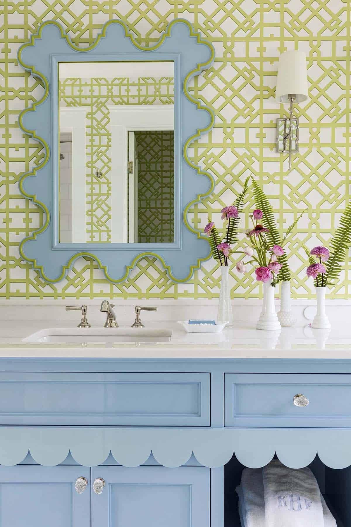 Scalloped blue mirror above bathroom sink with green trellis wallpaper and milk-glass bud vases