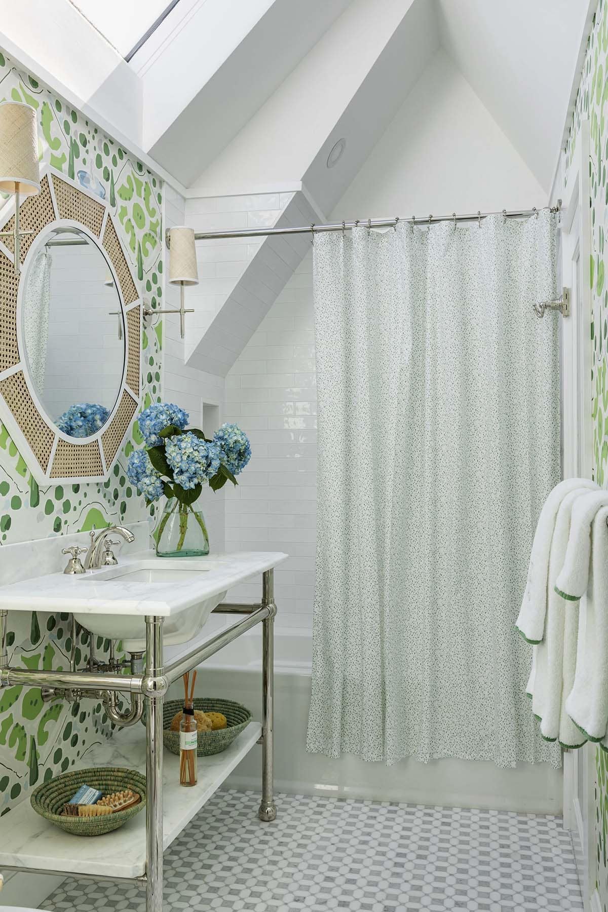 Skylit bathroom with green patterned wallpaper, cane mirror, and polka-dot shower curtain