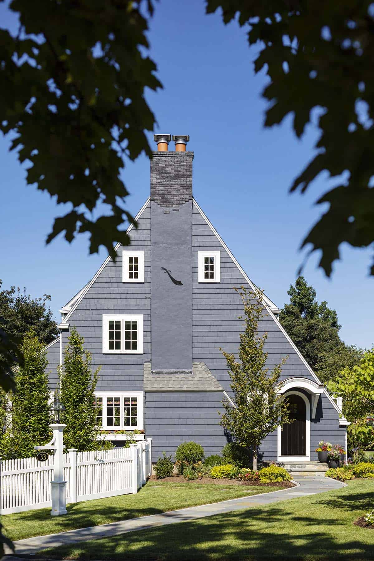 Gray shingle house exterior with steep gambrel roof, twin chimneys, and white picket fence