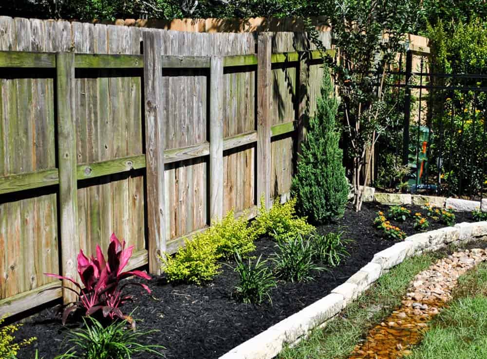 Curved stone-edged fenceline bed with junipers, shrubs, and a dry creek border in a Texas backyard