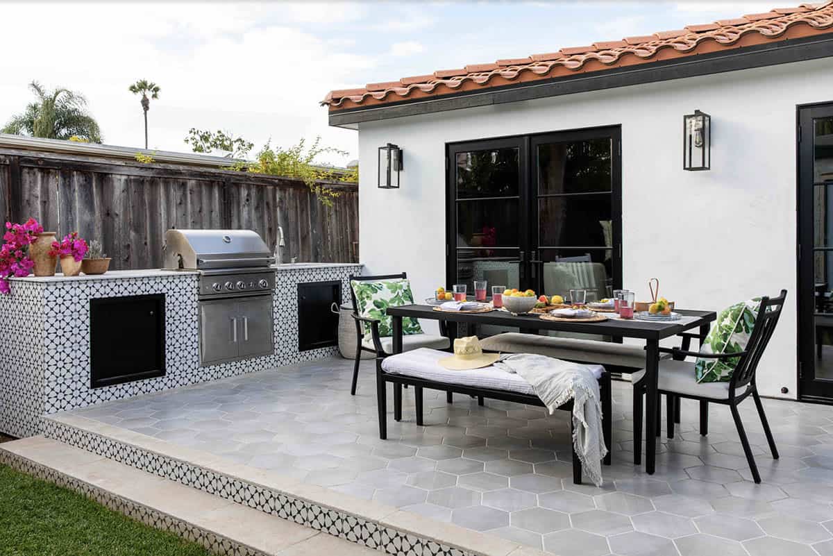 Spanish-style patio with a black and white patterned tile outdoor kitchen and built-in grill, next to a black dining table set with fruit and a bench topped with a striped throw and sun hat