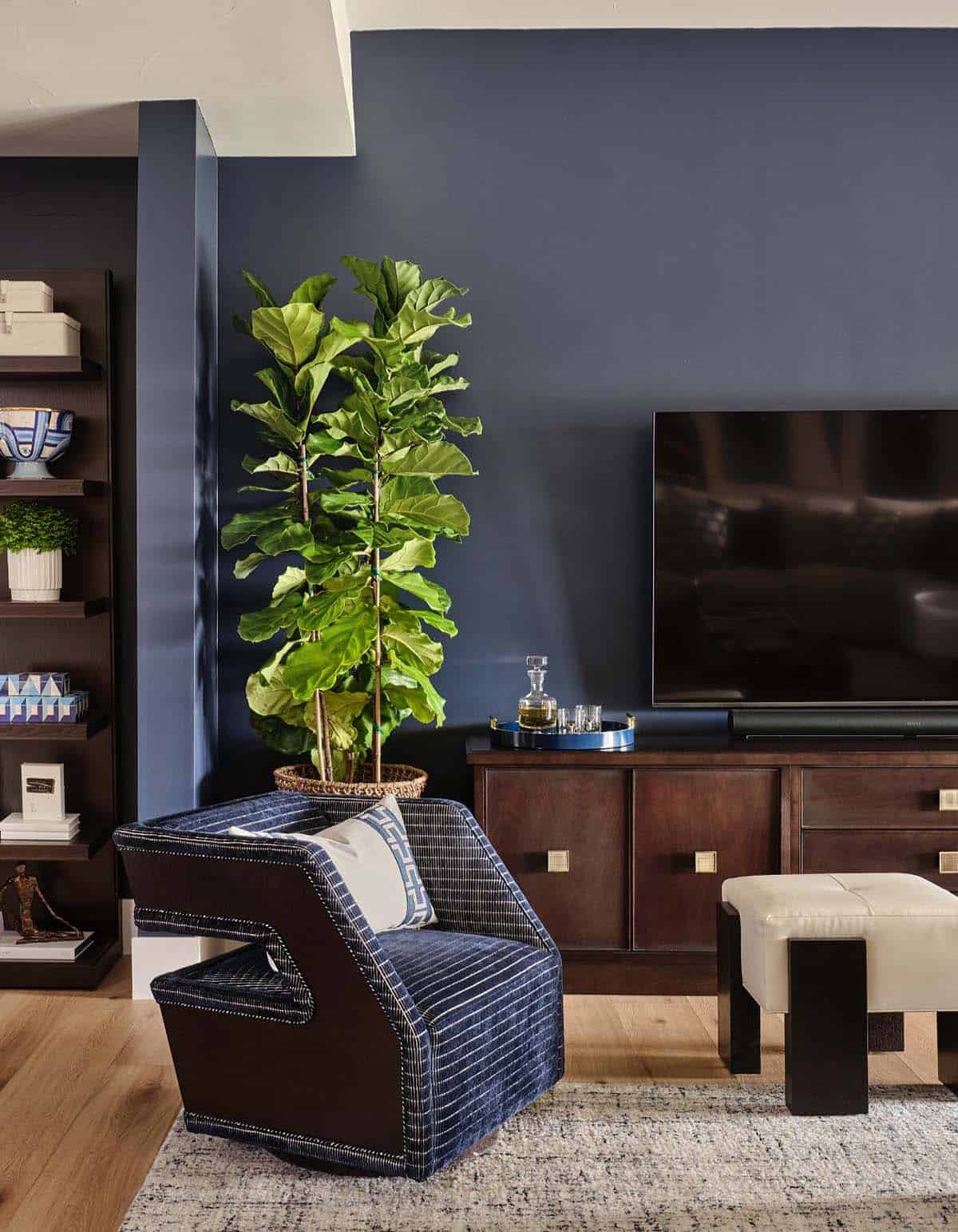 Navy accent wall with wall-mounted TV, dark wood media console, fiddle leaf fig plant, and striped blue armchair