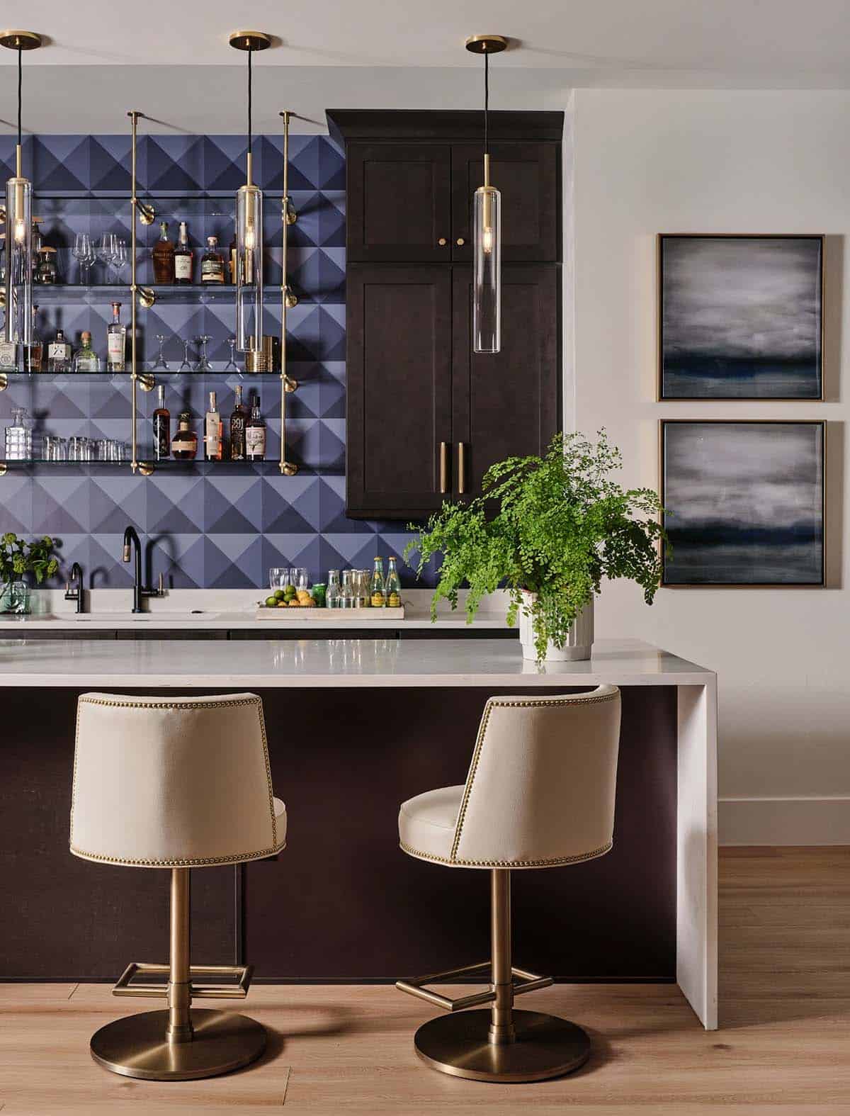 Home bar with geometric blue tile backsplash, glass shelving, brass pendant lights, and cream barstools