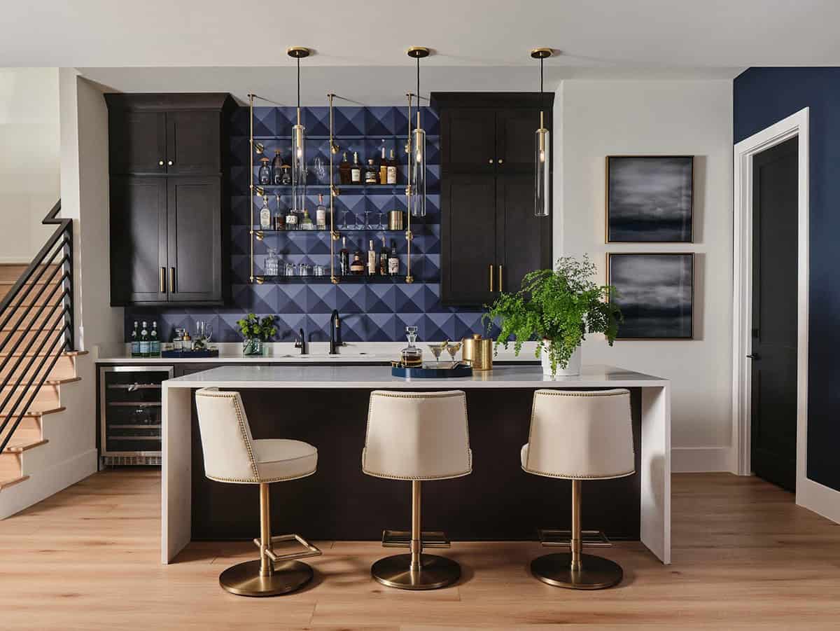 Wide view of home bar area with dark cabinetry, blue tile wall, glass bottle shelves, and three barstools at island