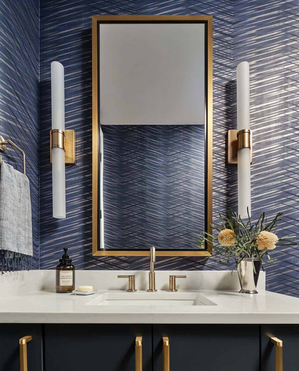 Powder room vanity with herringbone navy wallpaper, brass-framed mirror, gold sconces, and marble countertop