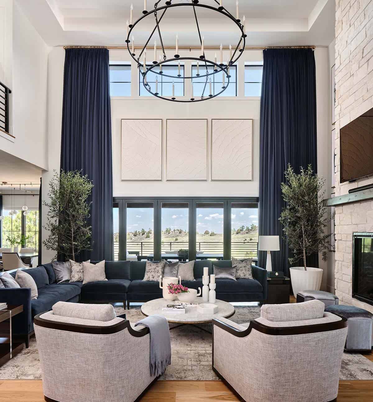 Two-story great room with navy velvet sectional, tall navy curtains, round chandelier, and floor-to-ceiling windows