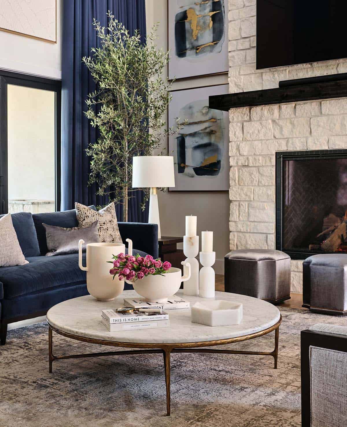 Round marble coffee table styled with ceramic vases, pink tulips, candle holders, near stone fireplace