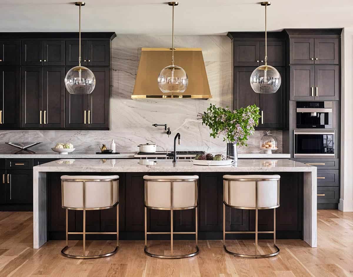 Kitchen island with dark wood cabinetry, brass hood, marble backsplash, glass pendant lights, and cream barstools