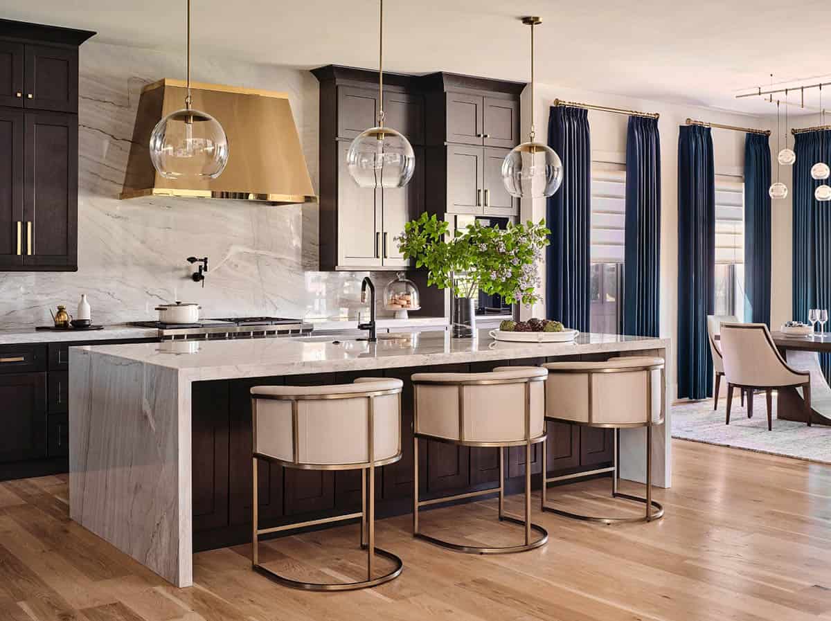 Kitchen with marble waterfall island, brass range hood, globe pendant lights, and view to navy-curtained dining area