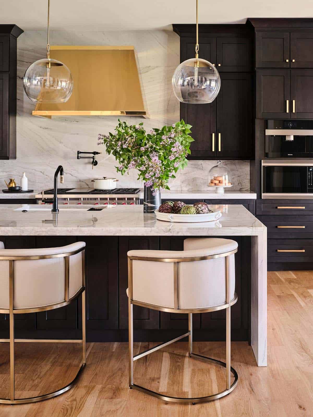 Close-up of kitchen island with brass hood, marble countertop, flowering branches, and cream-and-brass barstools