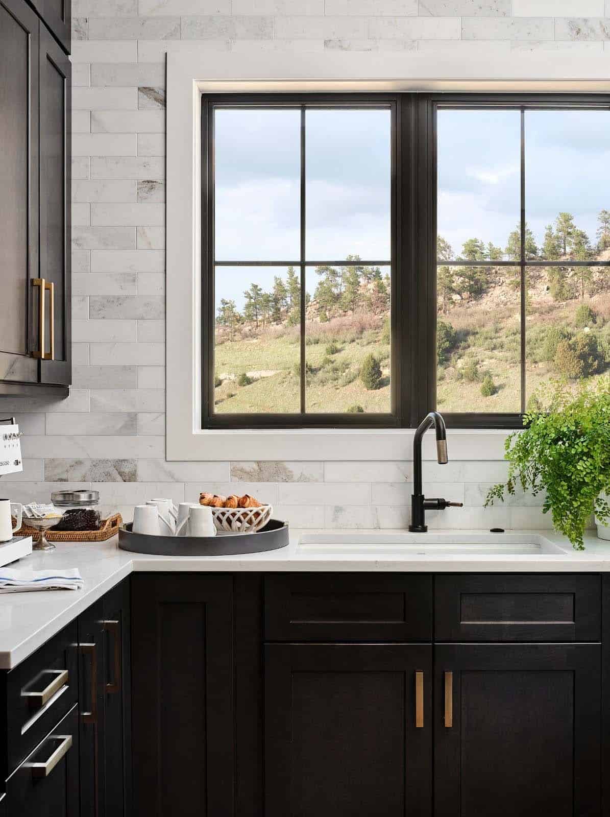 Kitchen sink area with marble subway tile backsplash, black-framed window overlooking hillside, and black faucet