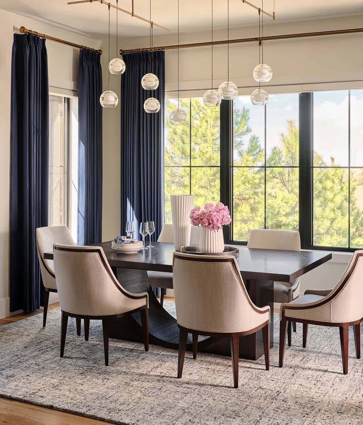 Dining table set with pink peonies, glassware, cream chairs, and linear brass pendant light fixture