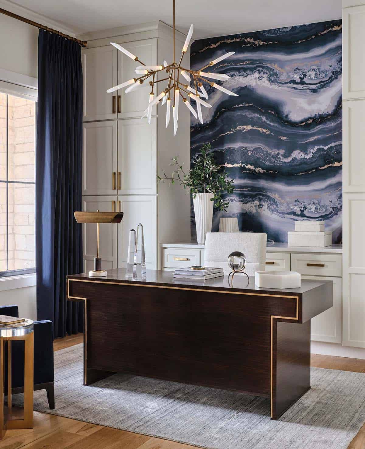 Home office with dark wood desk, gold trim, agate-patterned wall art, brass chandelier, and white cabinetry
