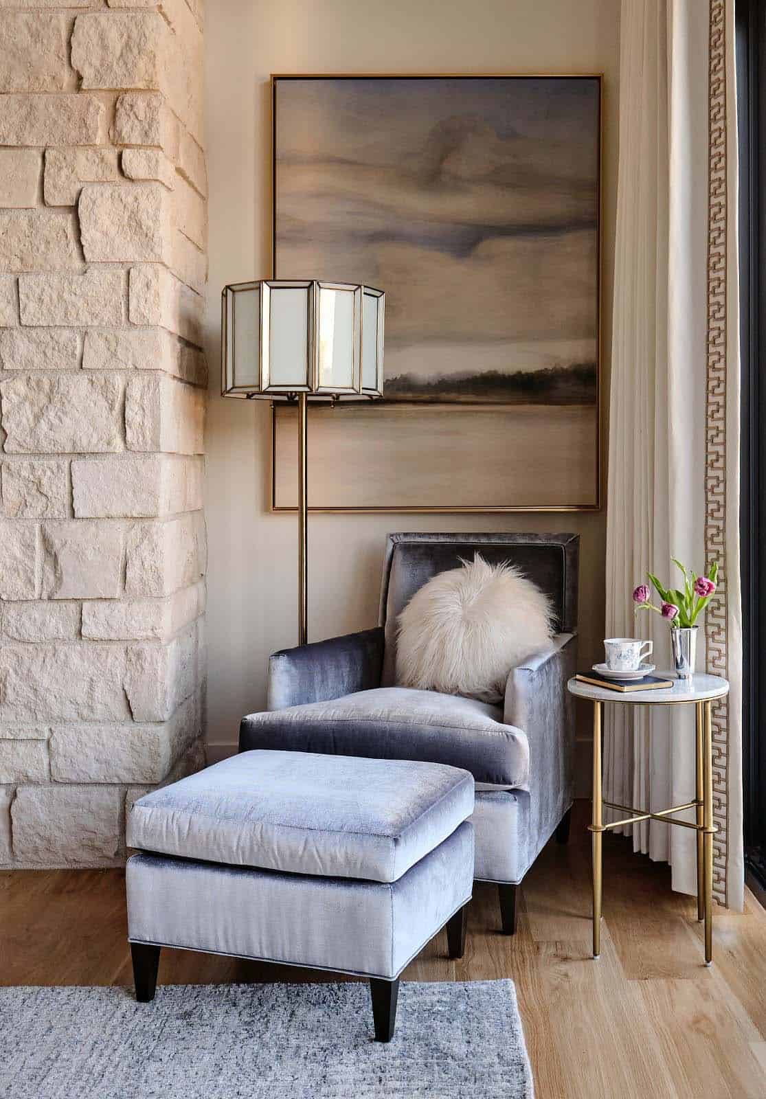 Reading nook with gray velvet armchair, ottoman, faux fur pillow, stone fireplace, and abstract landscape art