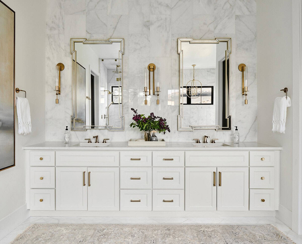 Double vanity bathroom with white cabinetry, marble tile wall, twin brass-framed mirrors, and gold sconces