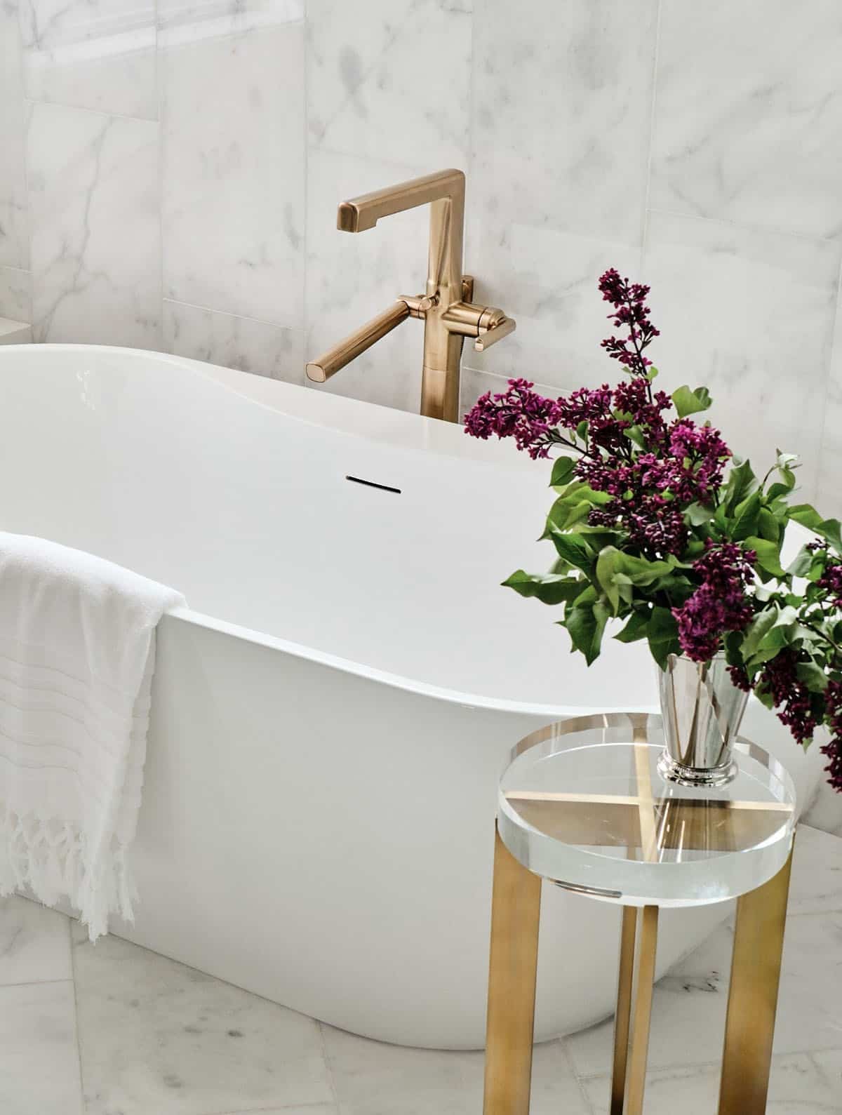 Close-up of white freestanding tub with brass filler faucet and lilac flowers on glass side table