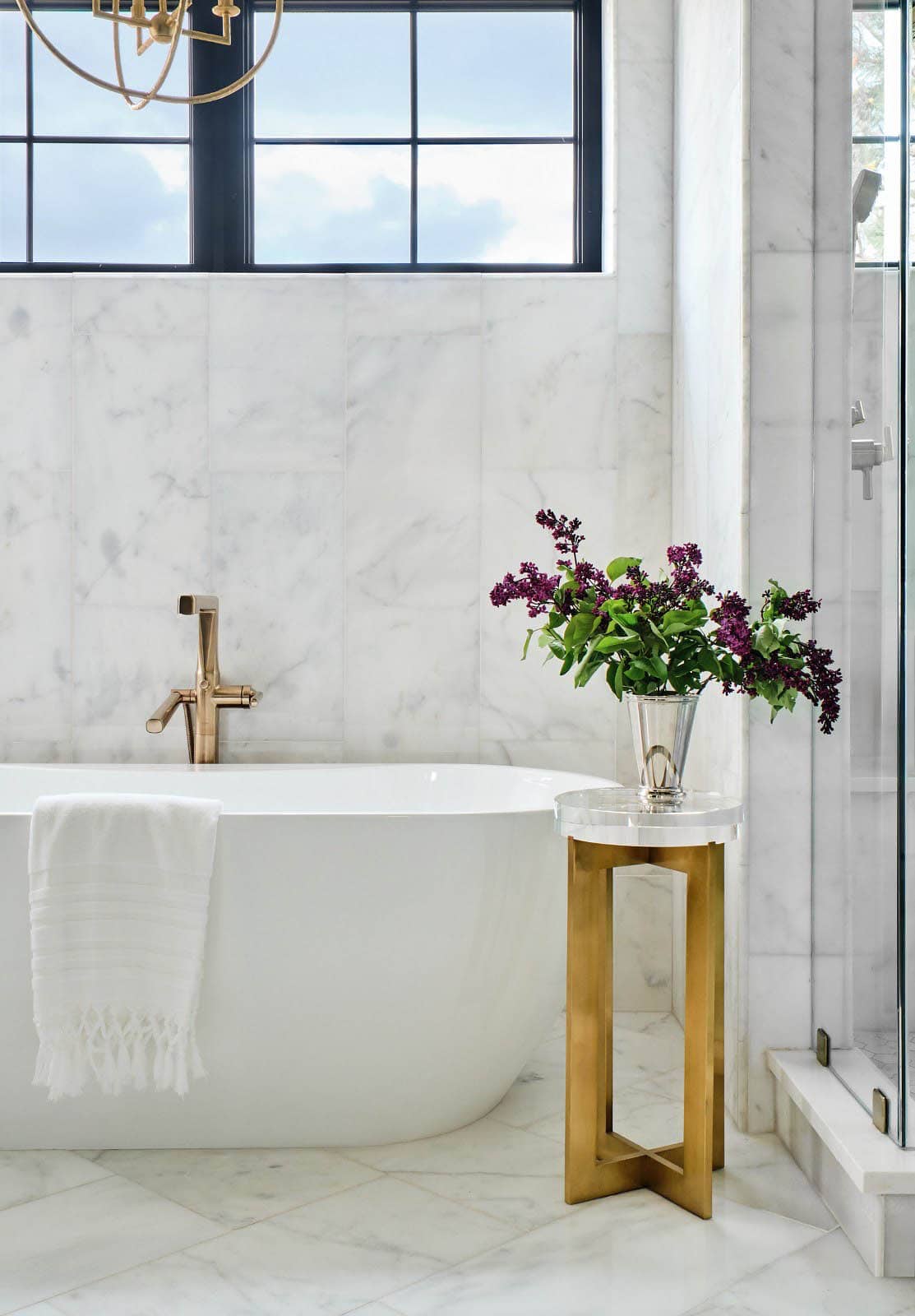 Freestanding tub beside glass shower, brass accent table with flowers, and marble tile walls under a window