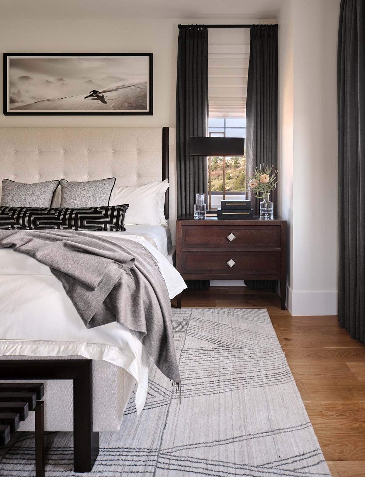 Bedside detail with dark wood nightstand, framed skiing photo, tufted headboard, and layered gray throw