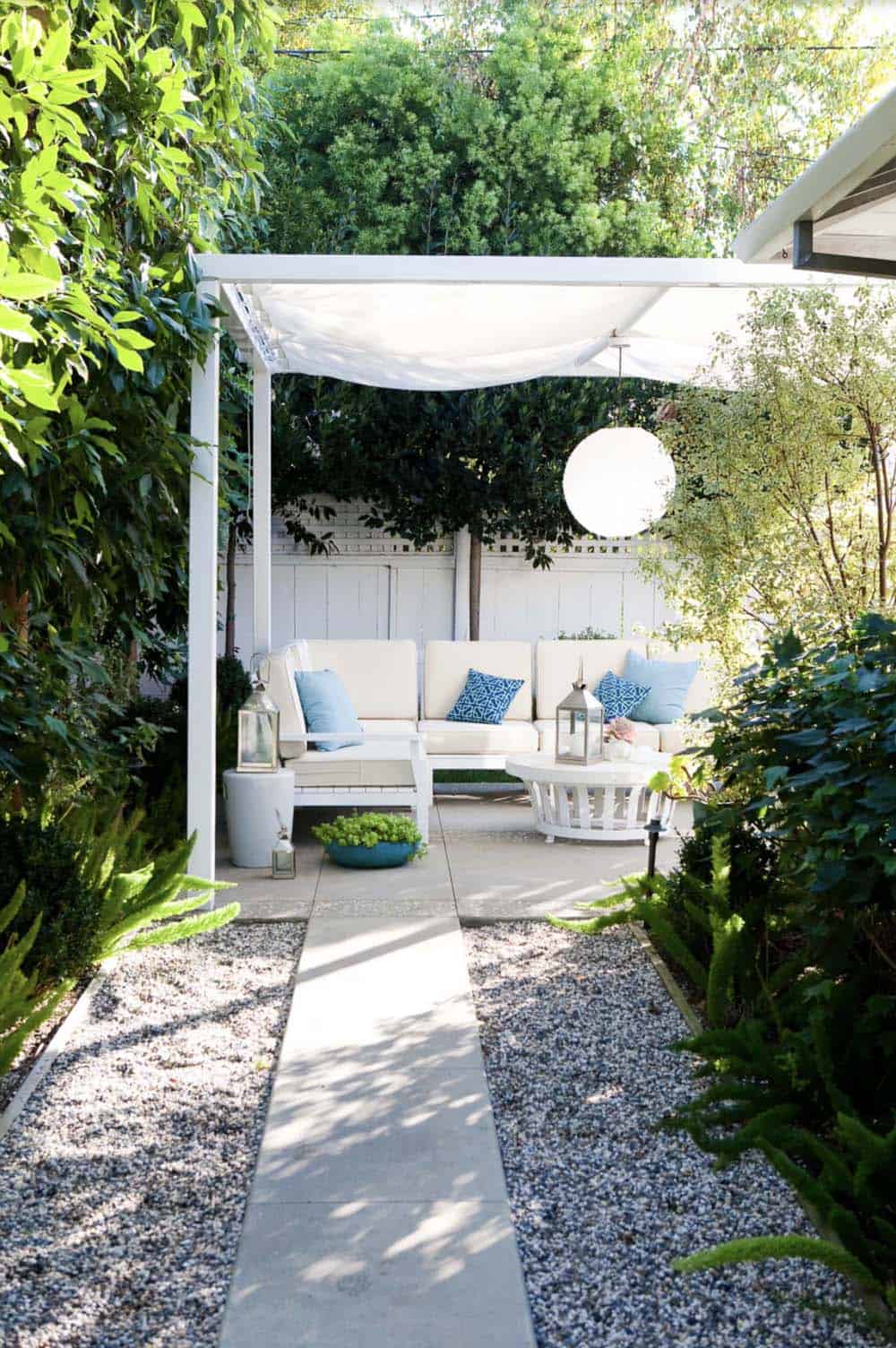 Tucked-away patio lounge with a white canvas canopy overhead, a white sectional with blue accent pillows, a round white side table, and a paper lantern light, surrounded by greenery