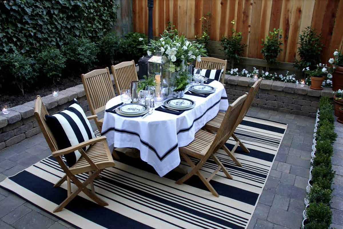 Formal outdoor dining table set with a white linen tablecloth, china, and candles, surrounded by teak folding chairs with black and white striped pillows, atop a matching striped outdoor rug