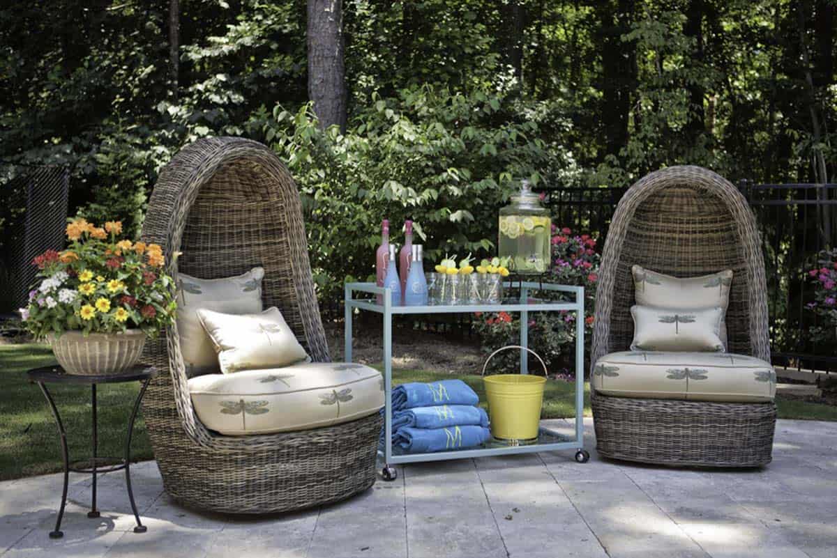 Two wicker egg chairs with dragonfly-print cushions flank a mint green rolling bar cart stocked with drinks, glasses, and rolled towels on a stone patio