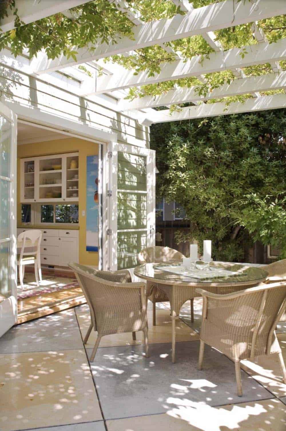 Outdoor wicker dining table and chairs set beneath a white pergola draped in wisteria, positioned just outside open French doors leading into a kitchen and dining room