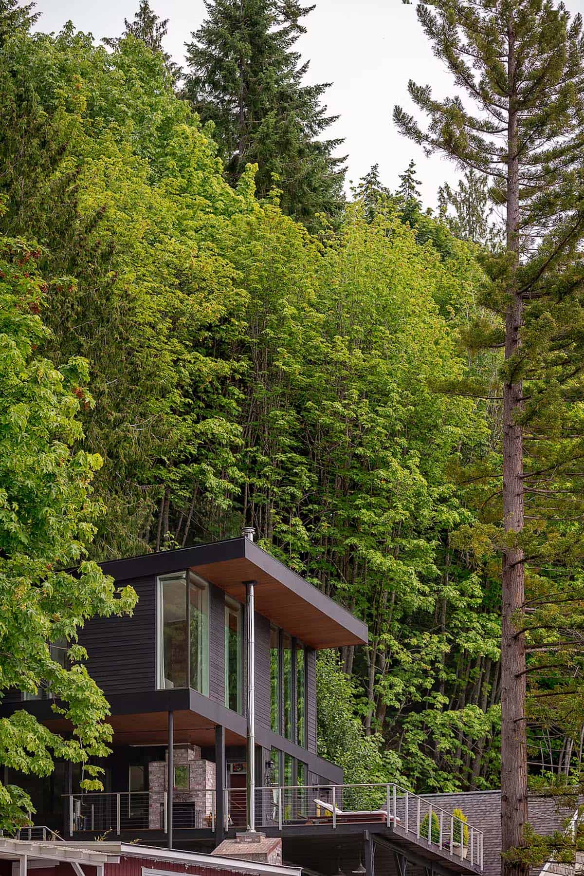 Black wood house with wide roof overhang and steel chimney flue, nestled against dense forest hillside