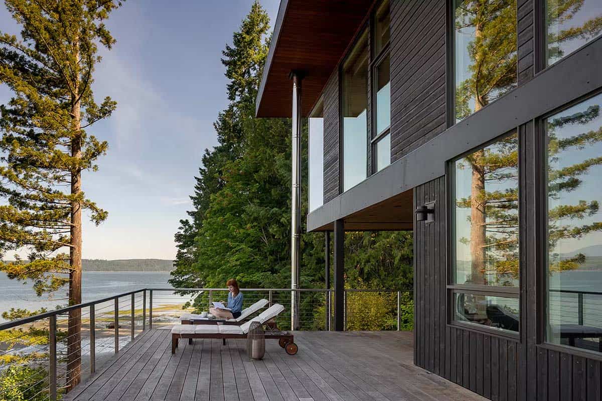 Woman reading on lounge chair on wooden deck beside dark timber-clad house, water and trees in view