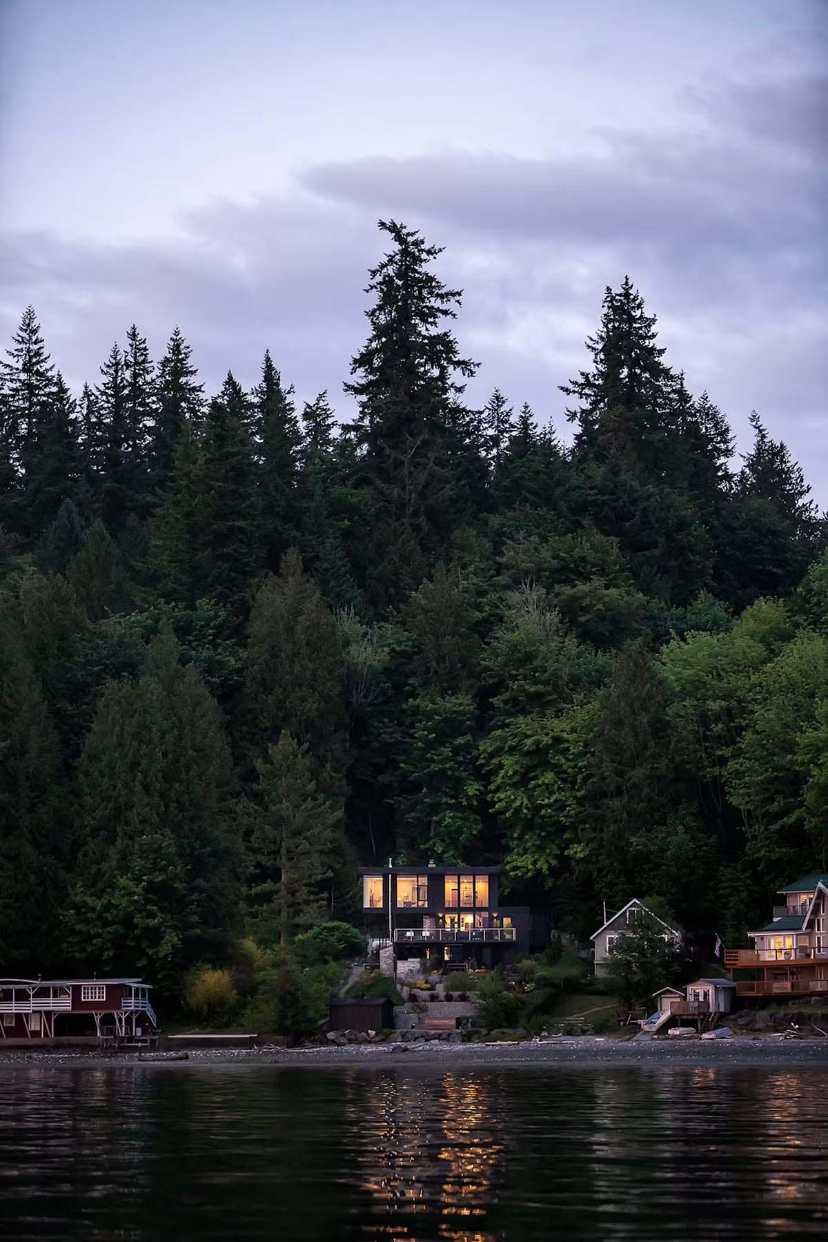 Illuminated house glowing at dusk on tree-covered shoreline, viewed from the water with neighboring cabins nearby