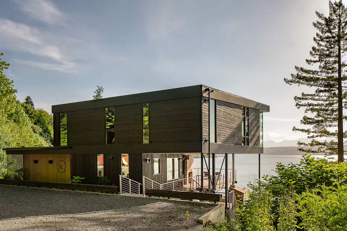 Elevated dark timber house on steel stilts above waterfront, gravel driveway and pine tree in foreground