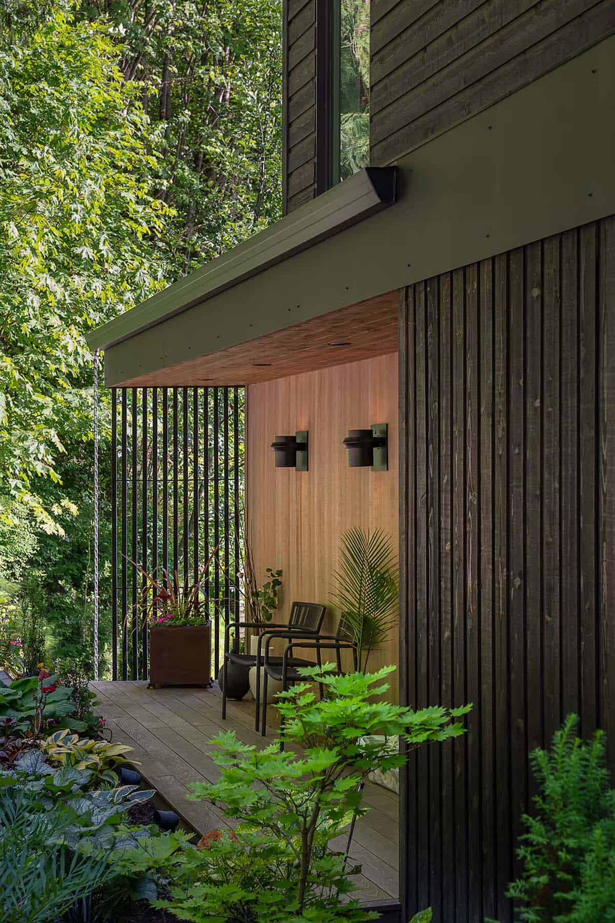 Covered outdoor entry nook with wood paneling, black chairs, and potted plants amid slatted wood screen