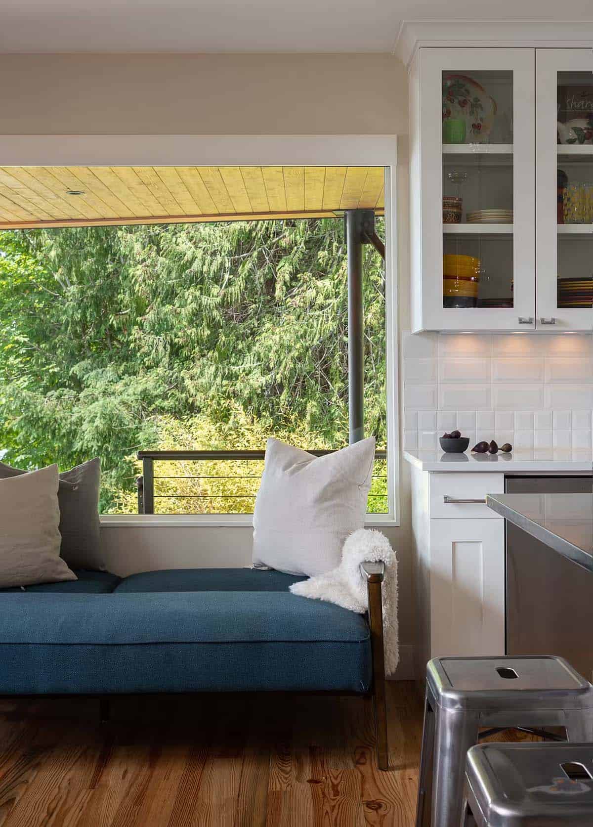 Blue daybed with pillows beside large window overlooking deck and trees, white kitchen cabinets nearby