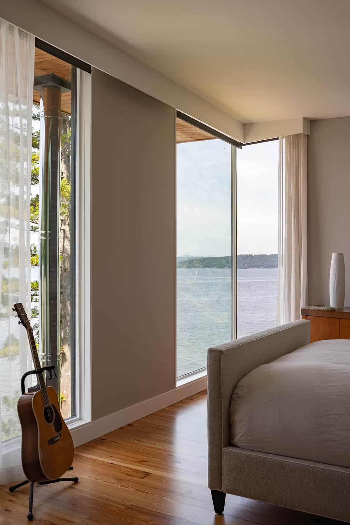 Bedroom with upholstered bed, acoustic guitar on stand, and floor-to-ceiling windows framing water view