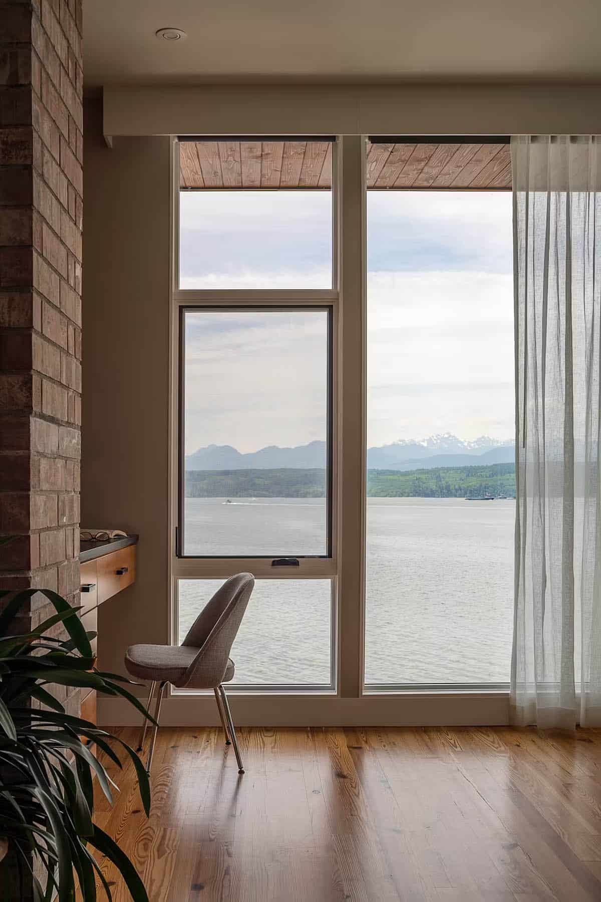 Reading nook with upholstered chair beside floor-to-ceiling windows overlooking water and distant snow-capped mountains