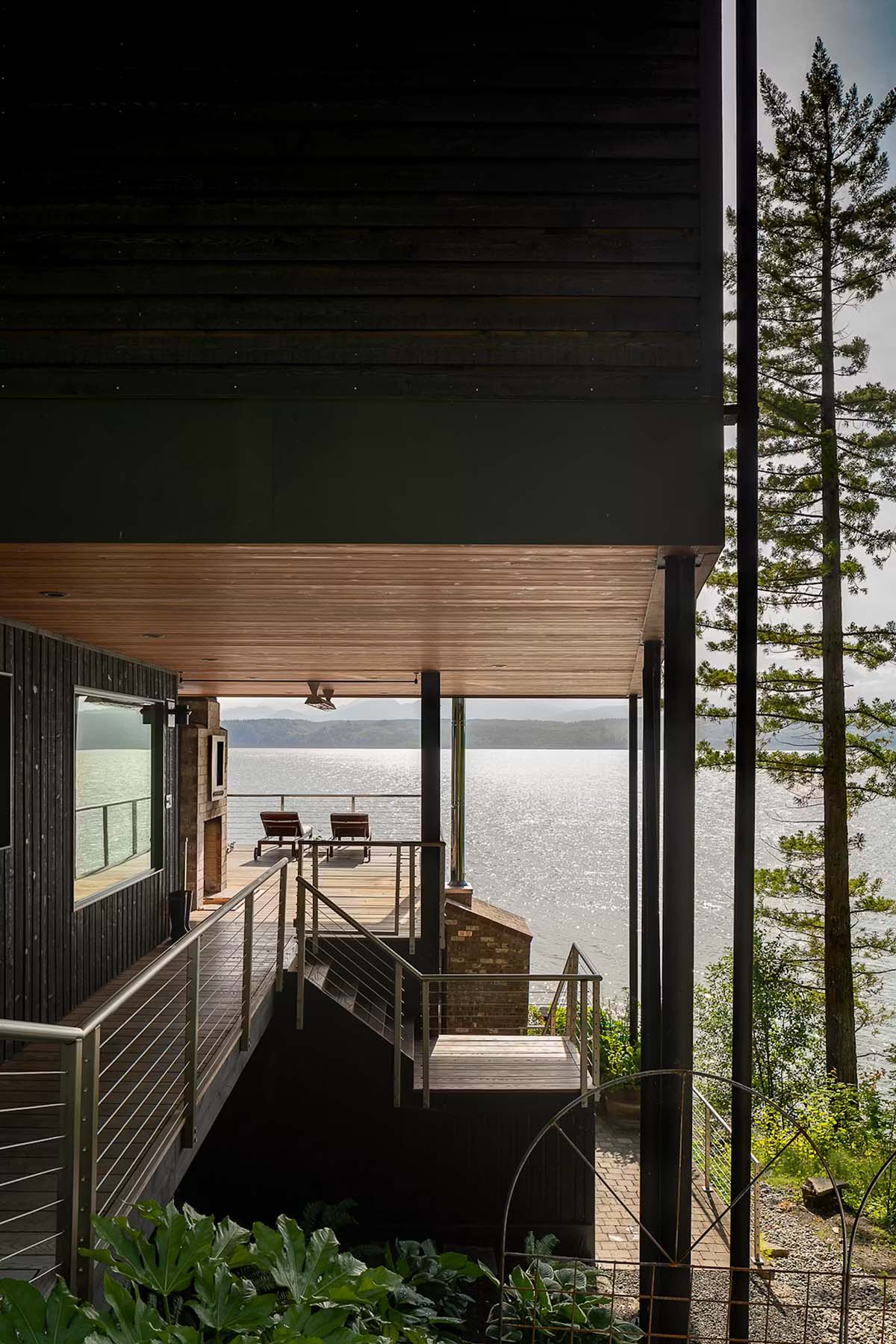 View from covered deck walkway toward lounge chairs and water, brick chimney and steel railings framing the scene