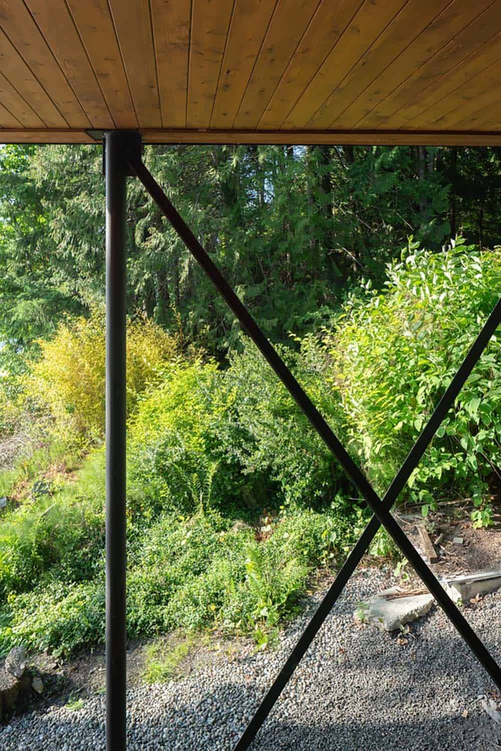 Steel X-brace support column beneath a wooden roof overhang, framing a lush garden view