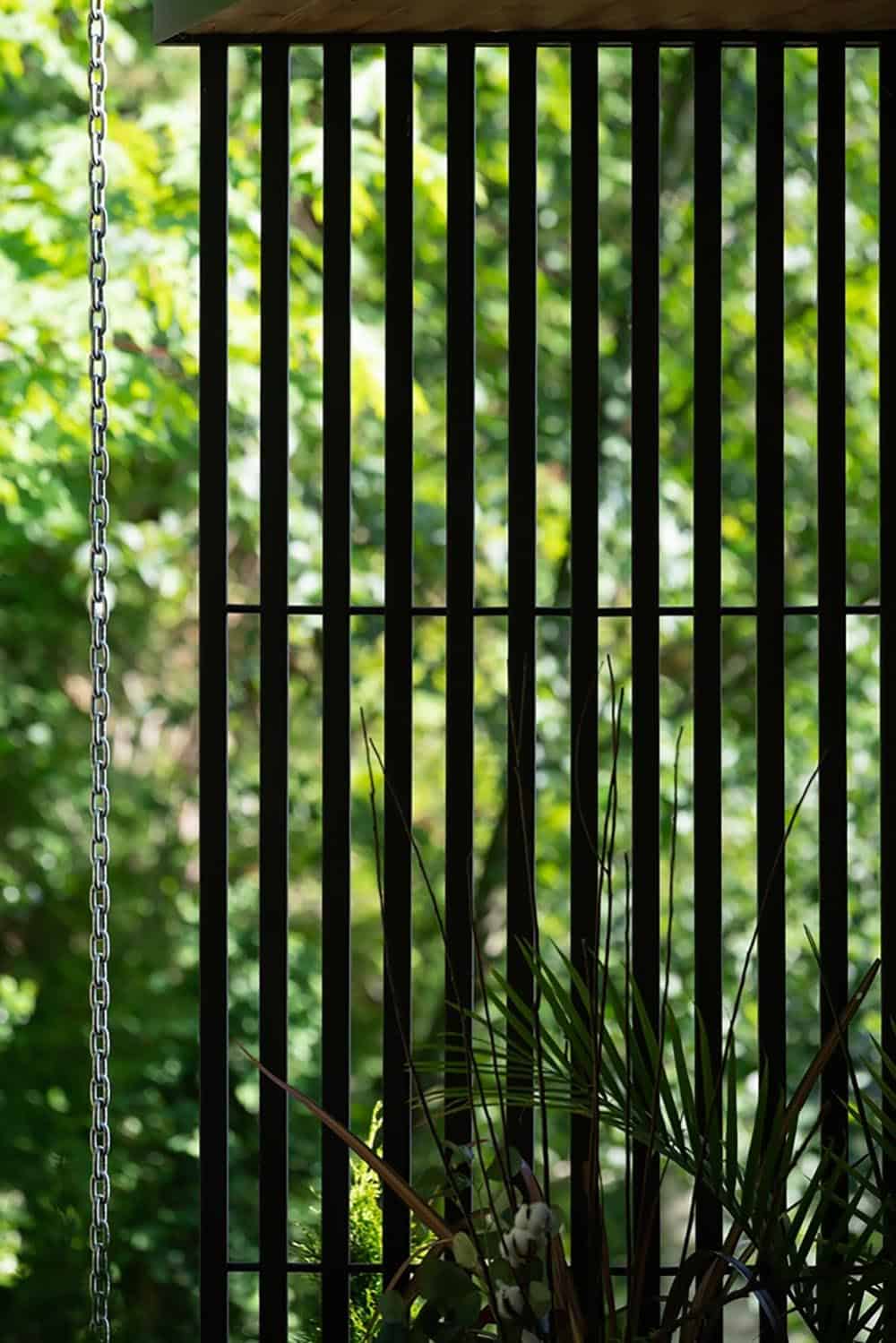 Vertical black wood slat screen with hanging chain, filtering light from a green garden