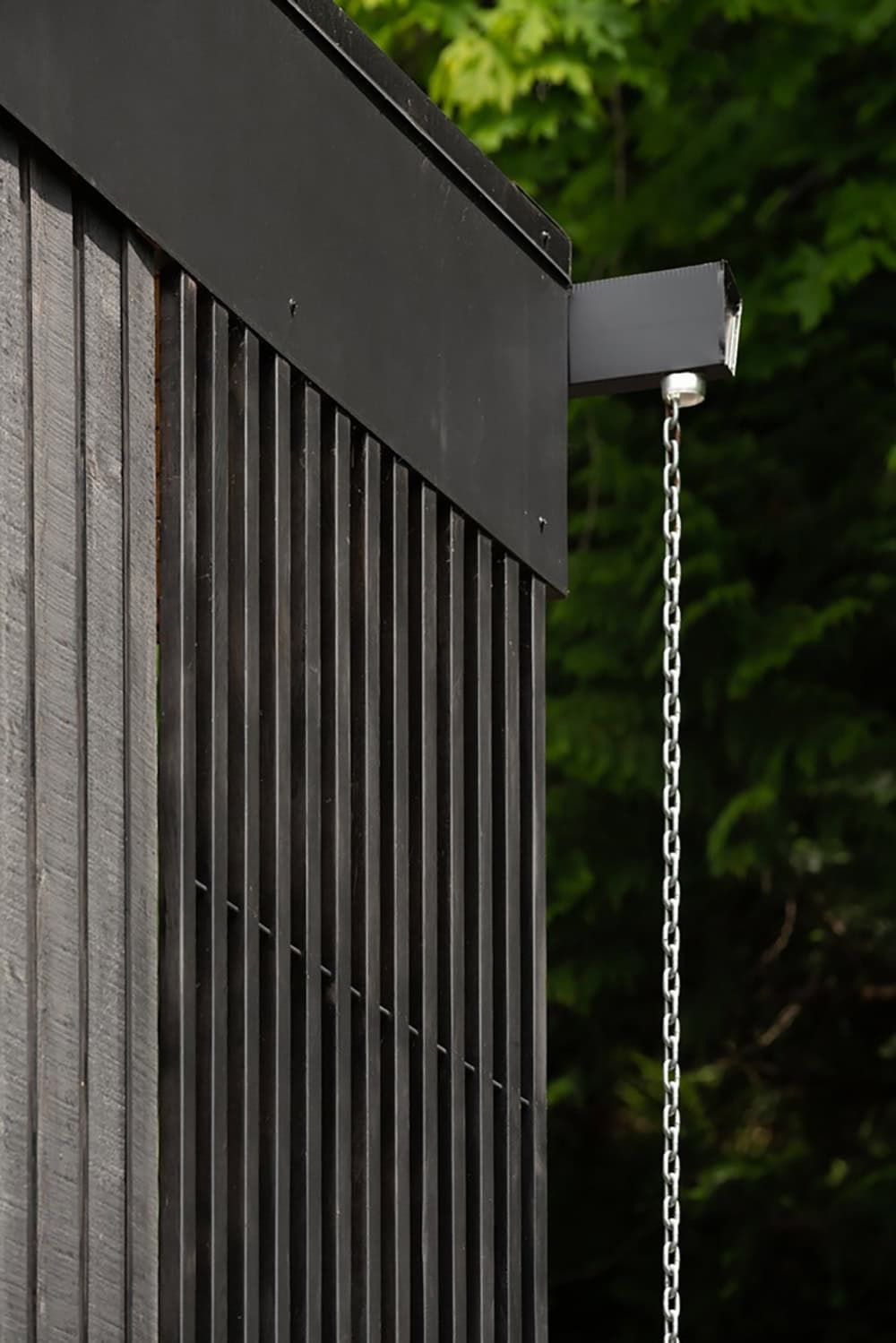 Corner detail of black wood siding and metal roof edge with rain chain against trees