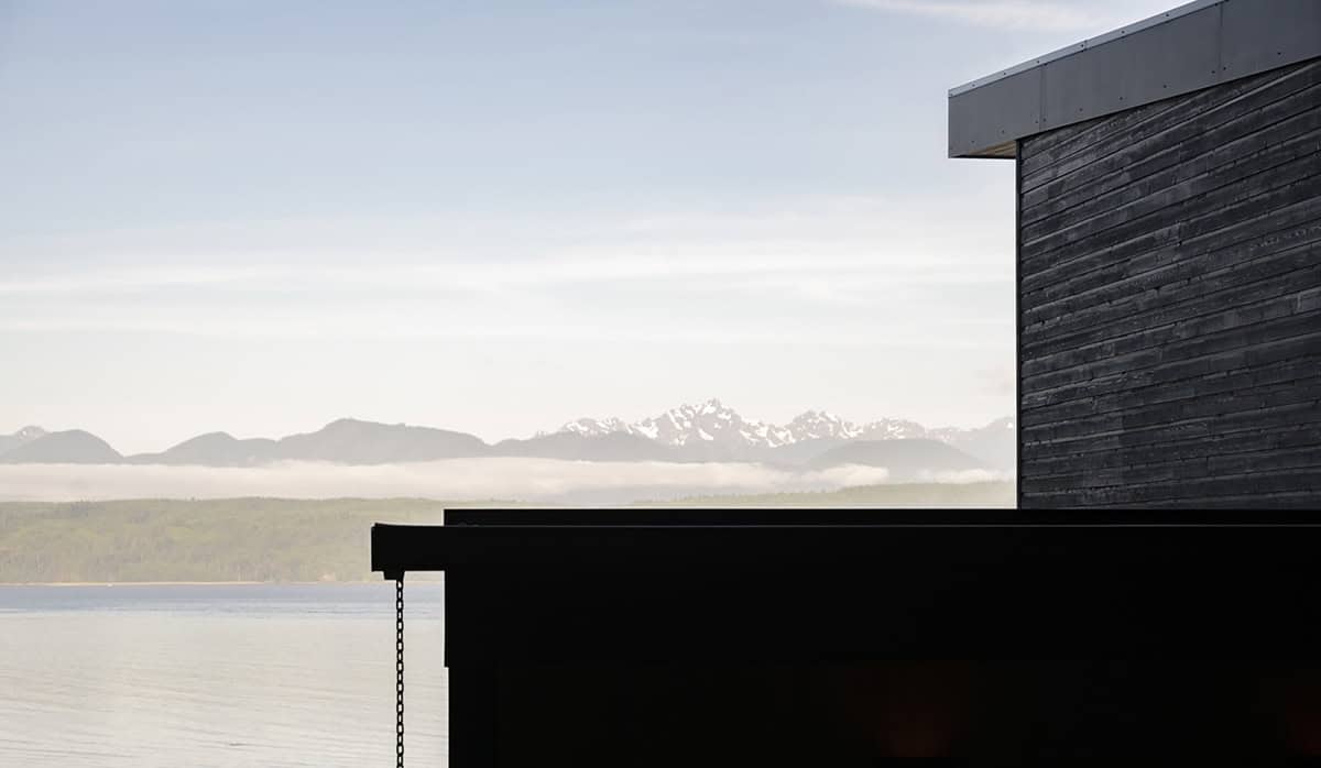 Silhouette of the black-clad home's roofline with distant snow-capped mountains and water beyond