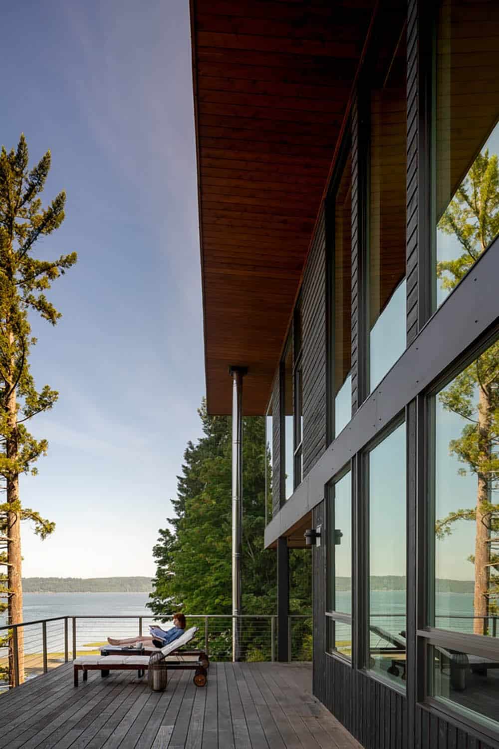 View along a deck under a wood-lined overhang with glass railing and water in the distance