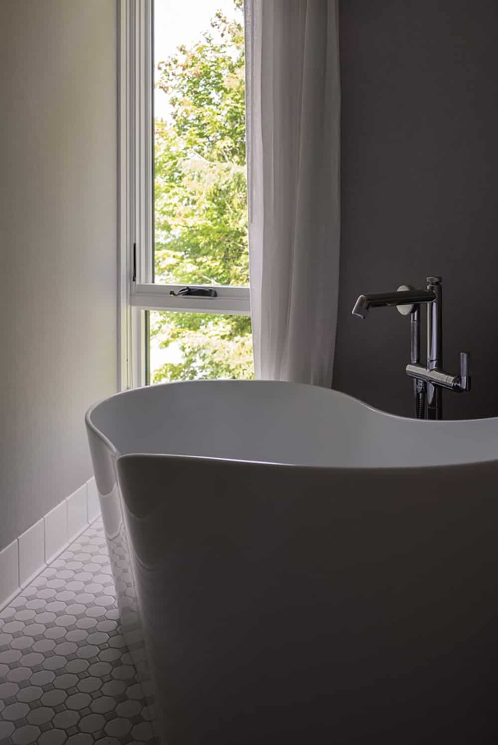 Freestanding bathtub with modern faucet beside a curtained window overlooking green trees