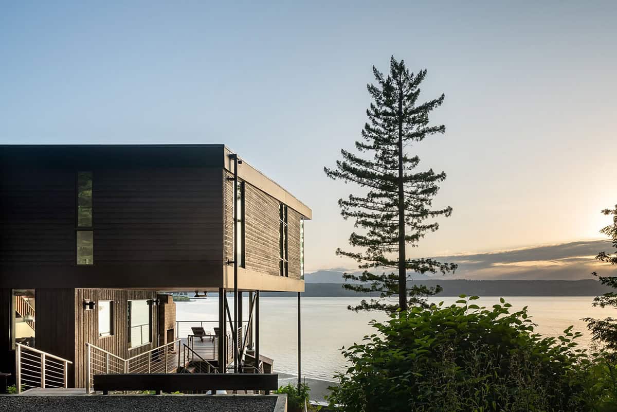 Dark cantilevered house beside a tall pine tree overlooking calm water at sunset, distant mountains on the horizon