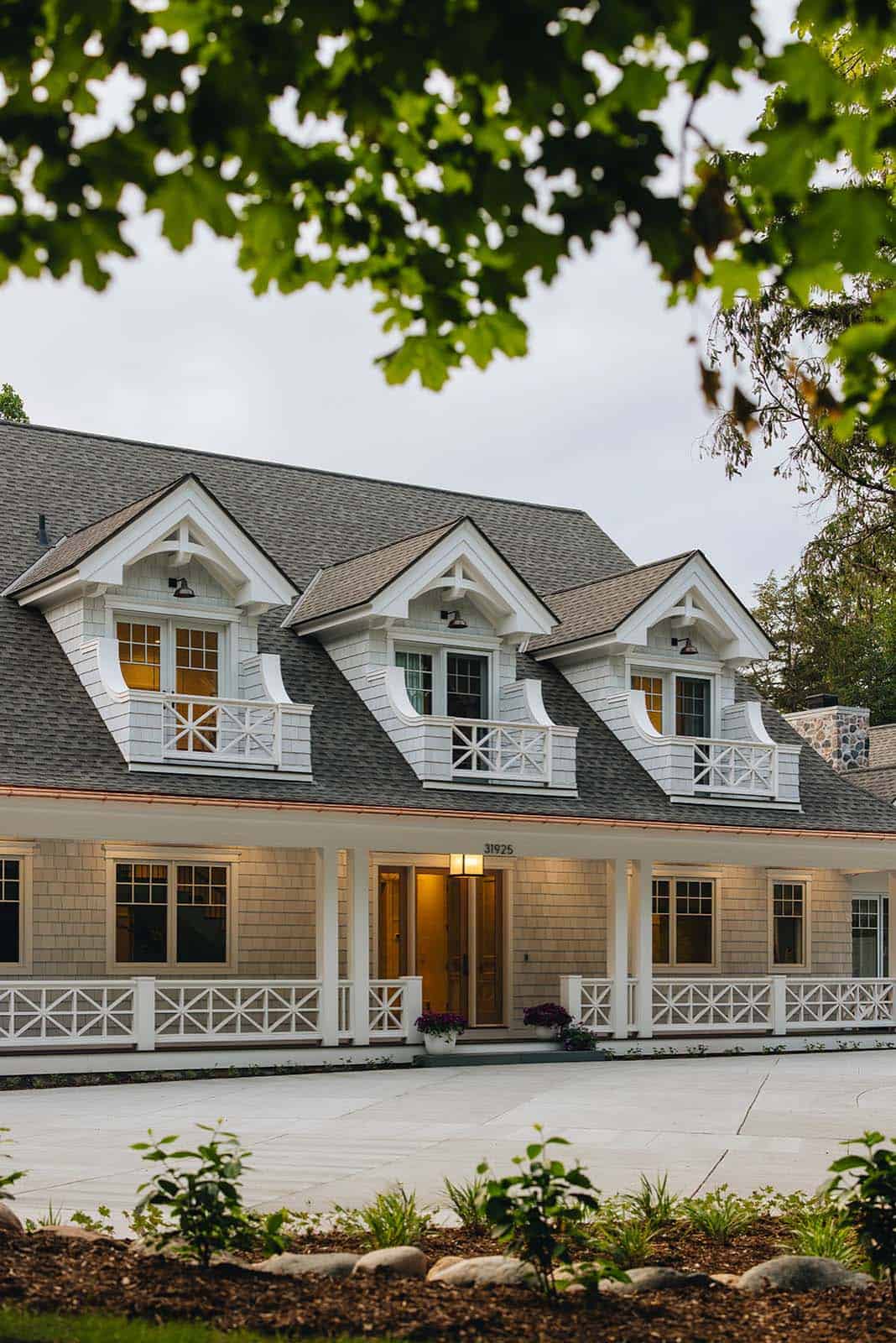 Front exterior of shingle-style lake house with three dormers framed by tree branches at dusk