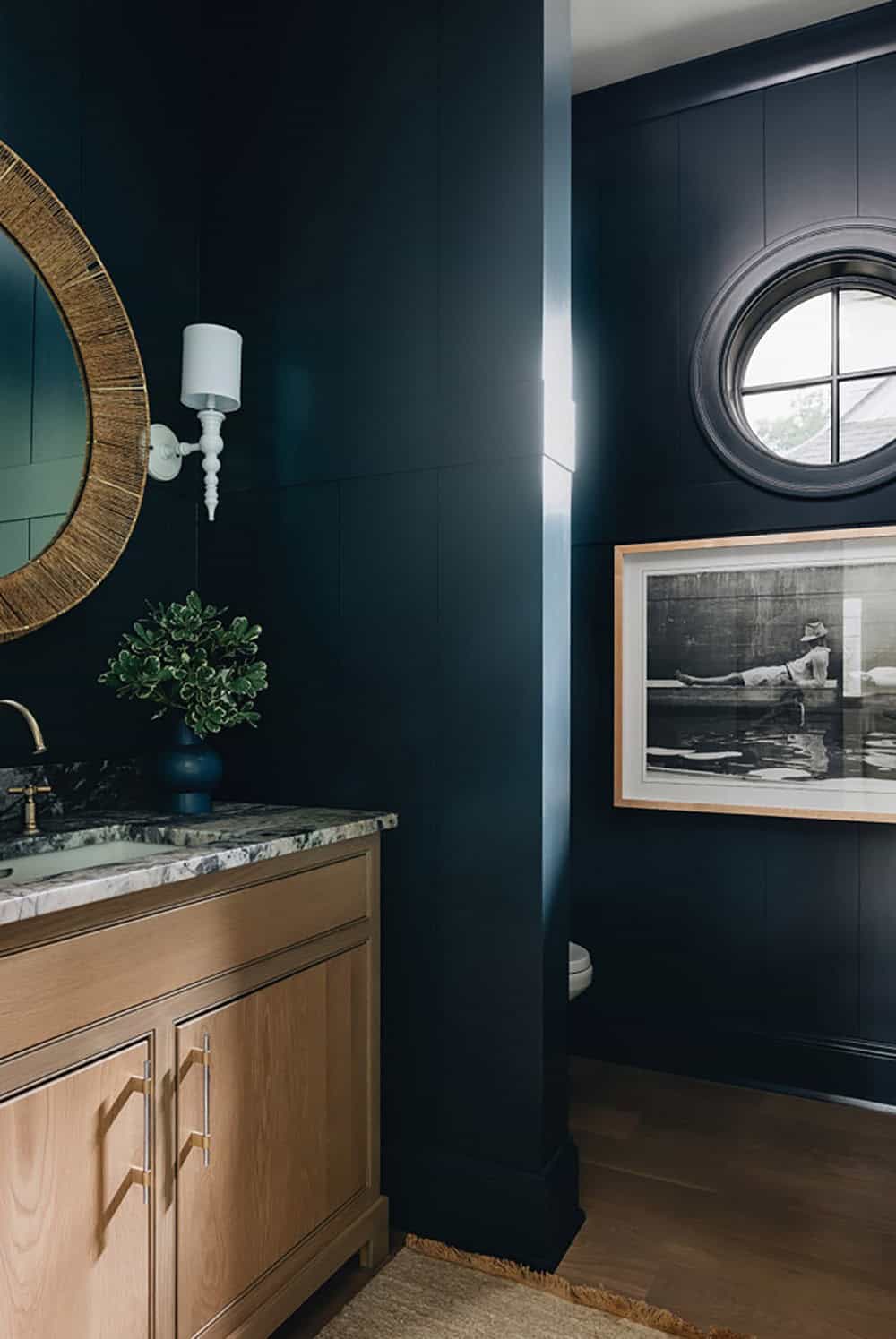 Navy powder room with round window, framed art, and marble vanity