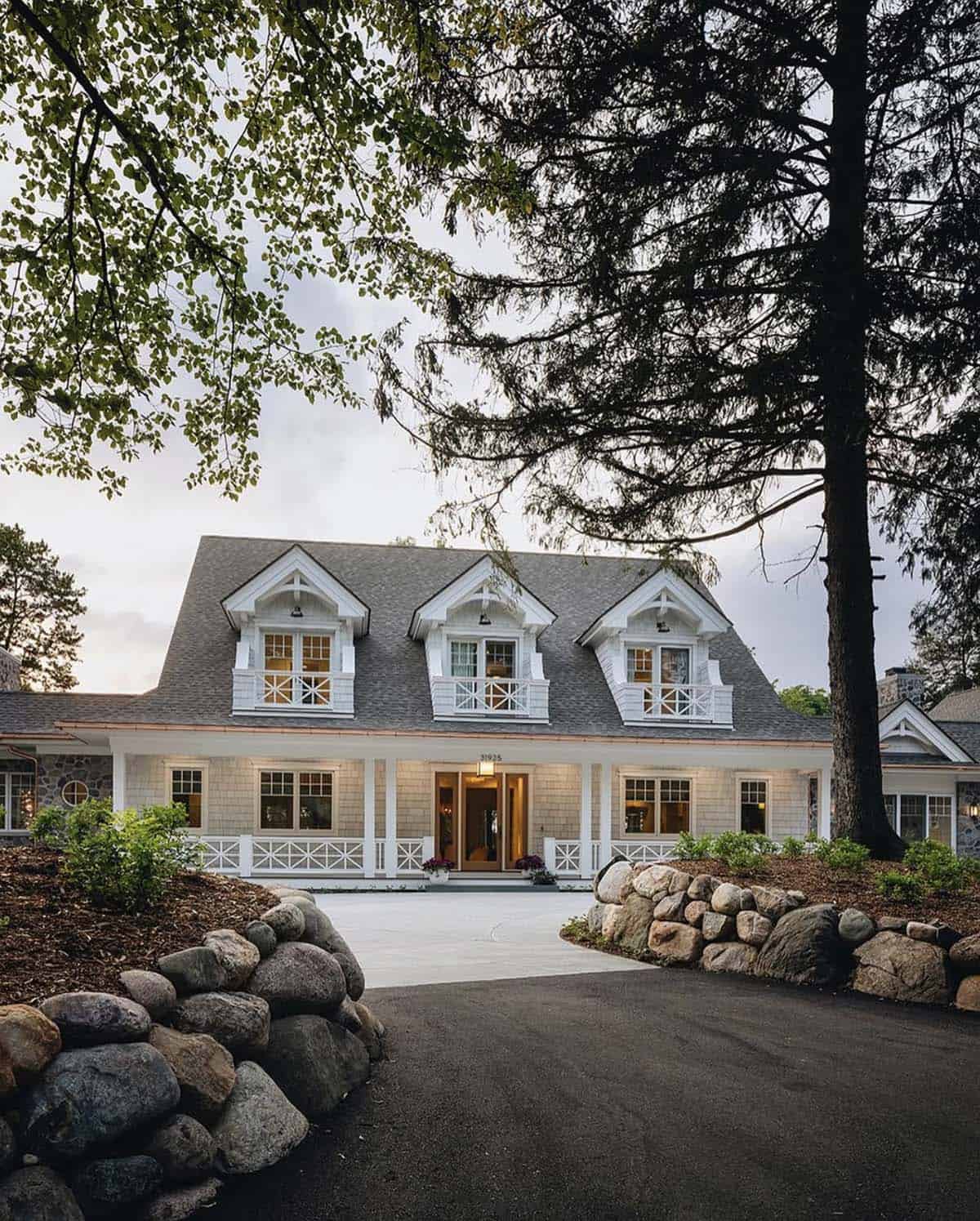 Front driveway view of shingle-style lake house framed by stone walls and trees at dusk